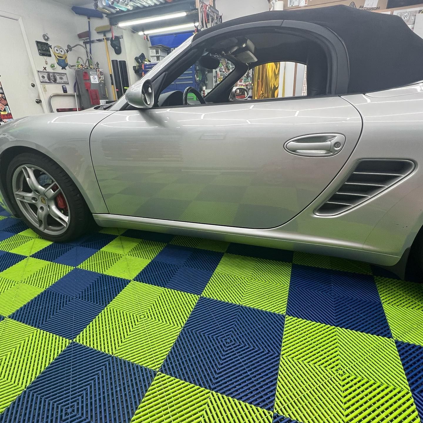 Silver Porsche Boxster convertible parked on blue and green checkered flooring in a garage.