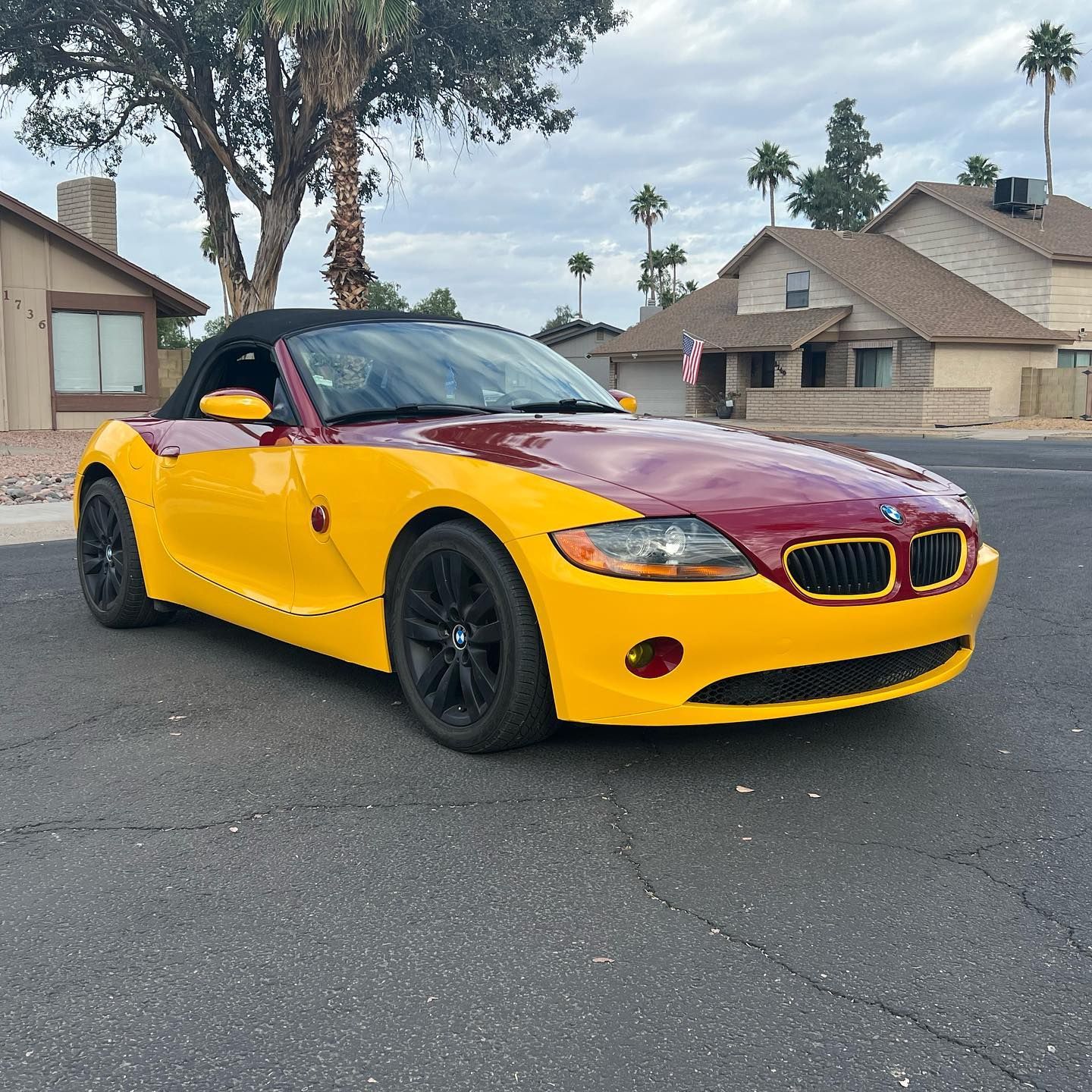 Yellow and maroon BMW Z4 convertible on a road in front of houses under a cloudy sky.