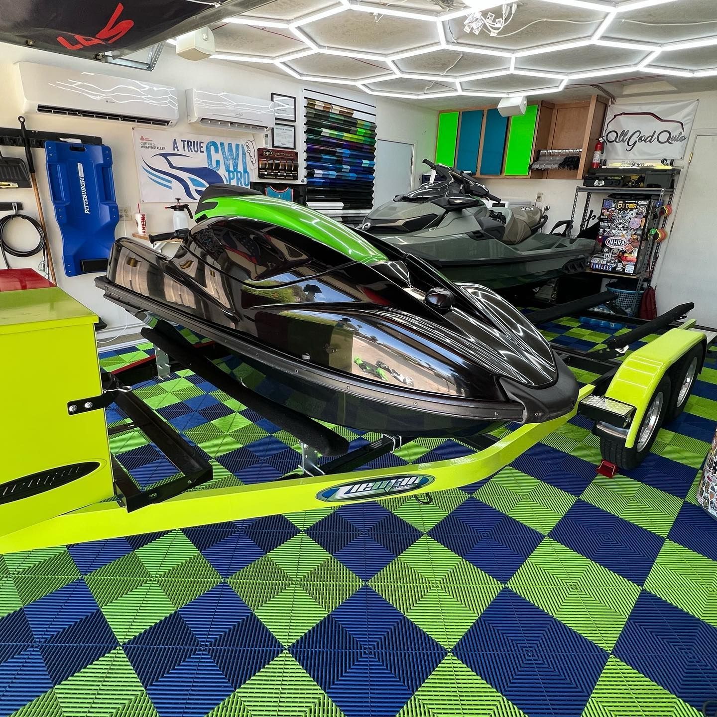 Black jet ski on a neon green trailer inside a garage with blue and green checkered flooring and a green jet ski.