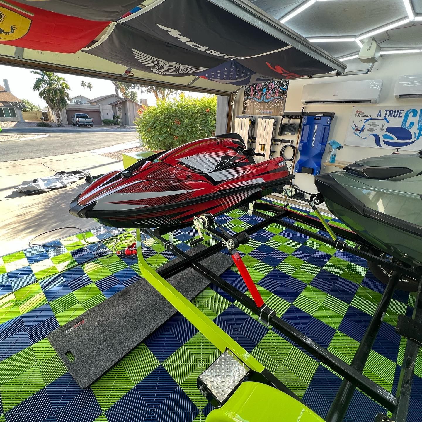 Red and gray jet ski on a green, blue, and black checkered platform inside a garage.