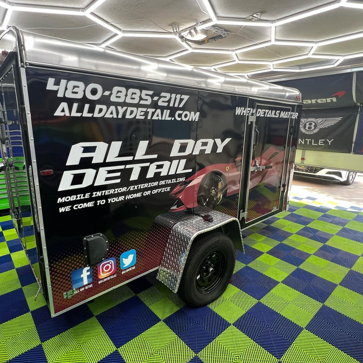 Black mobile detailing trailer with logo and contact info; parked inside a garage.