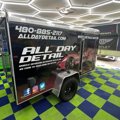 Black mobile detailing trailer with logo and contact info; parked inside a garage.