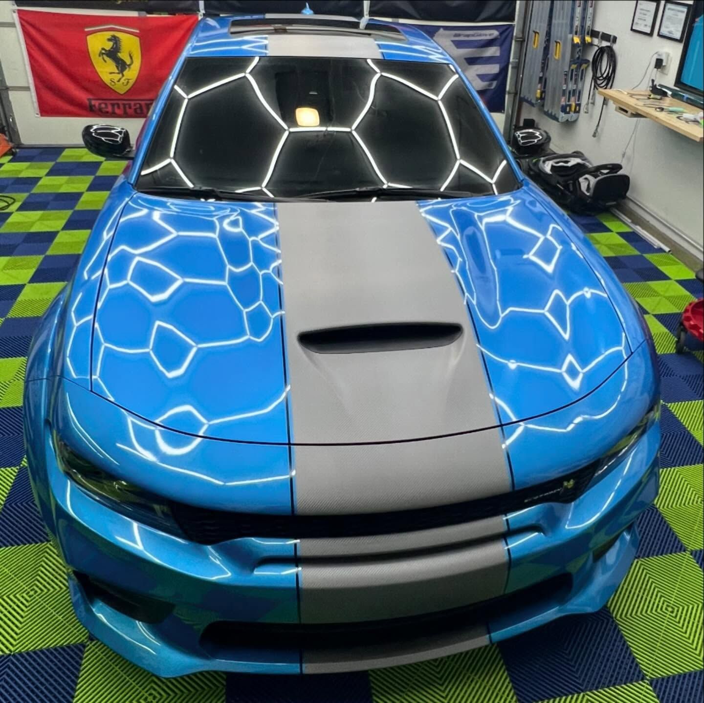 Blue and gray Dodge Charger with racing stripes in a garage with checkerboard flooring and a Ferrari flag.