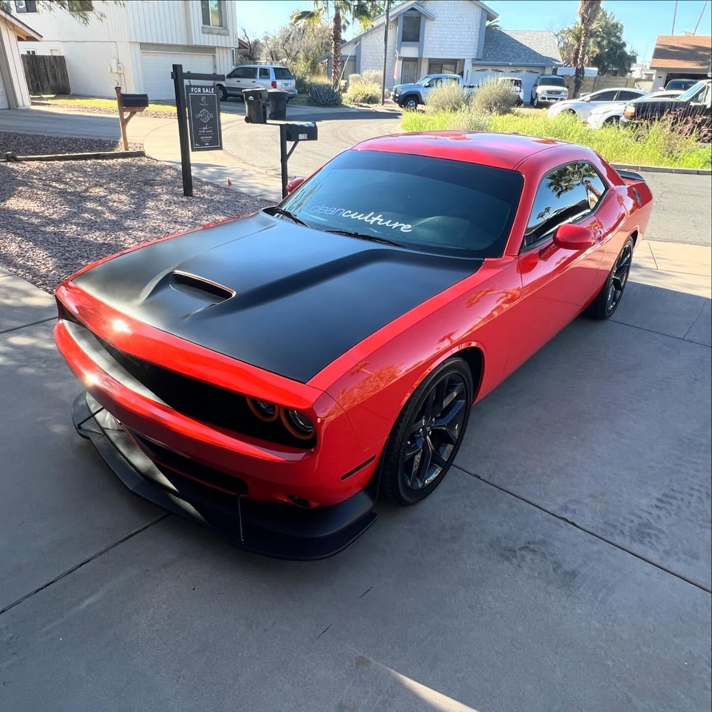 Red Dodge Challenger with a black hood parked on a driveway in front of houses on a sunny day.