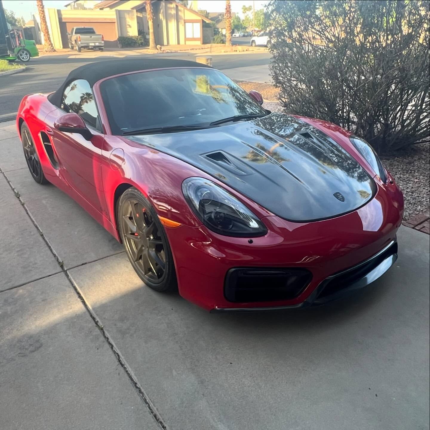 Red Porsche Boxster convertible with a black hood parked on a driveway in front of a house.