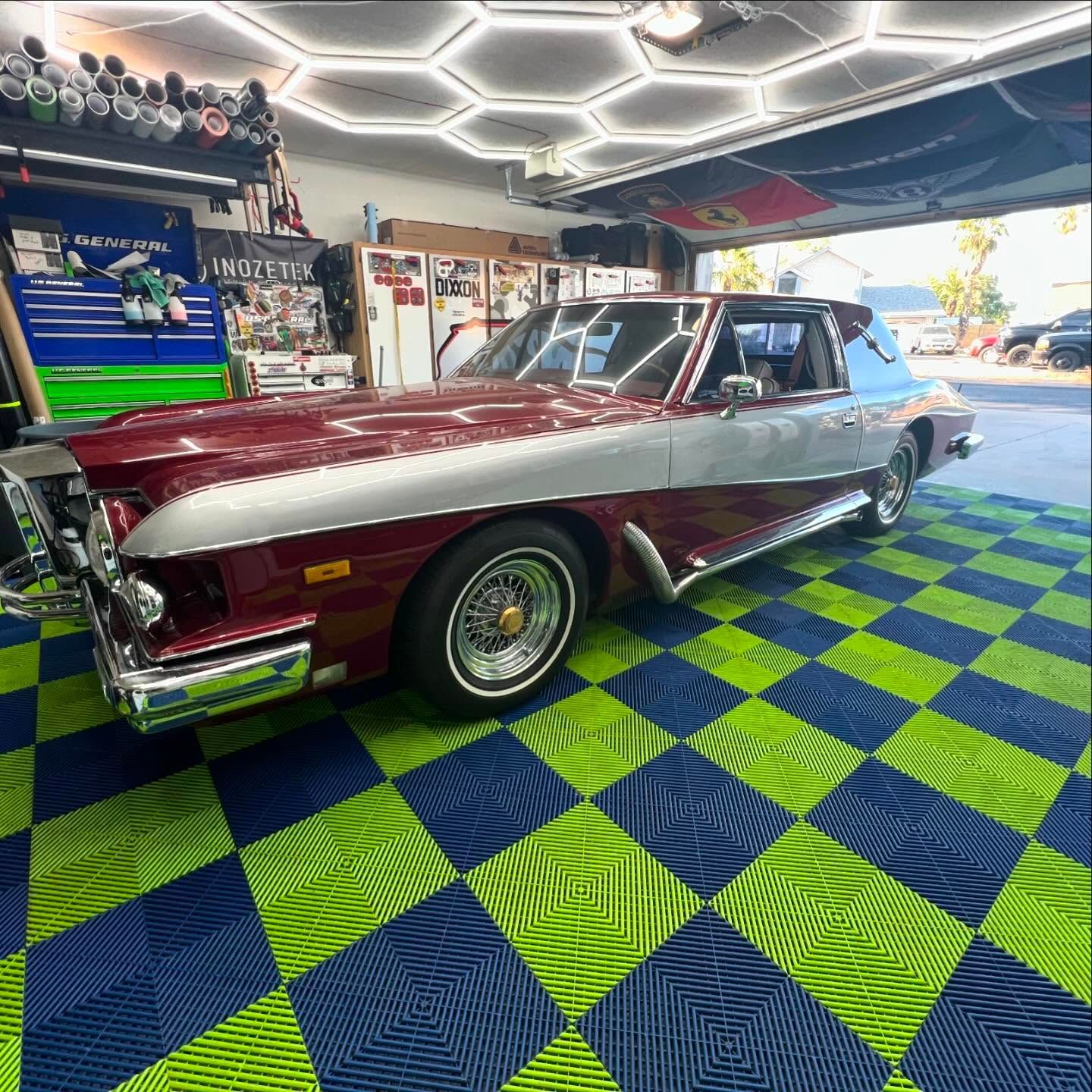 Classic burgundy and silver car parked in a garage with blue and green checkered flooring.