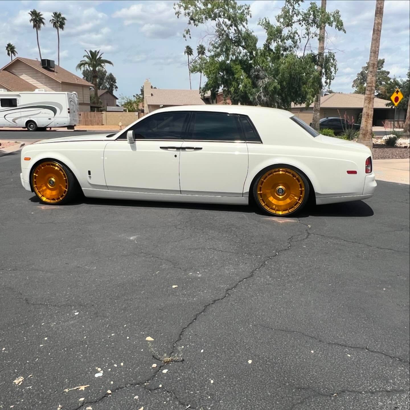 White Rolls-Royce with gold rims parked on asphalt in front of a residential area.