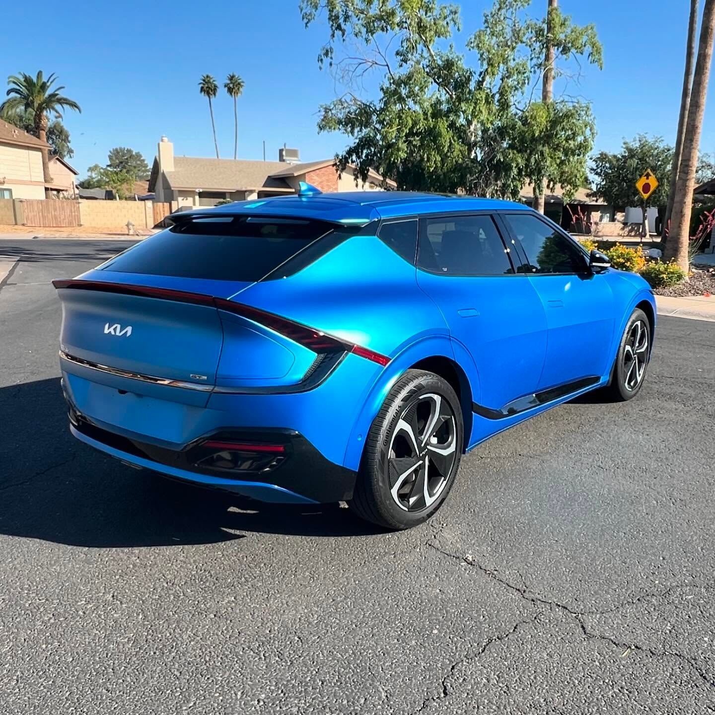 Blue Kia EV6 electric SUV parked on a paved street in a residential neighborhood under a sunny sky.
