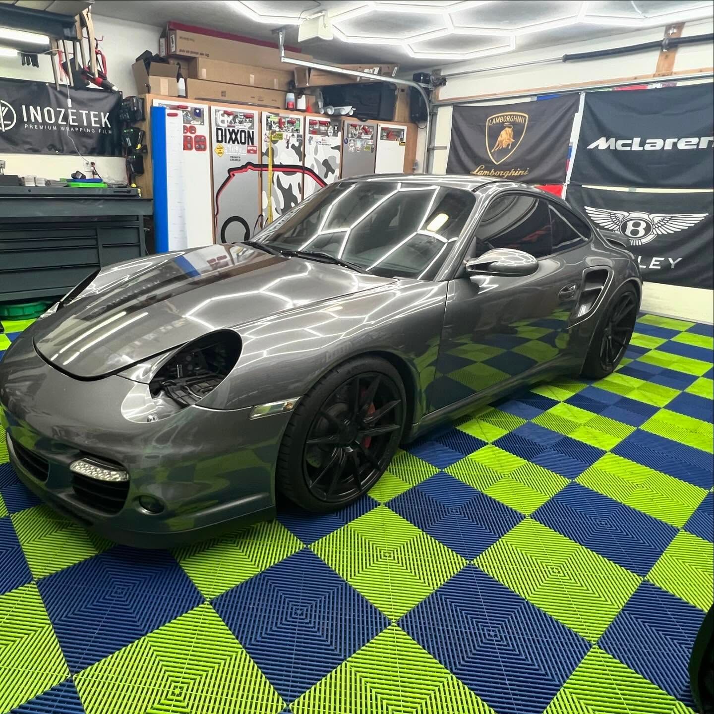 Gray Porsche sports car in a garage with blue/green checkered floor, under bright lights.