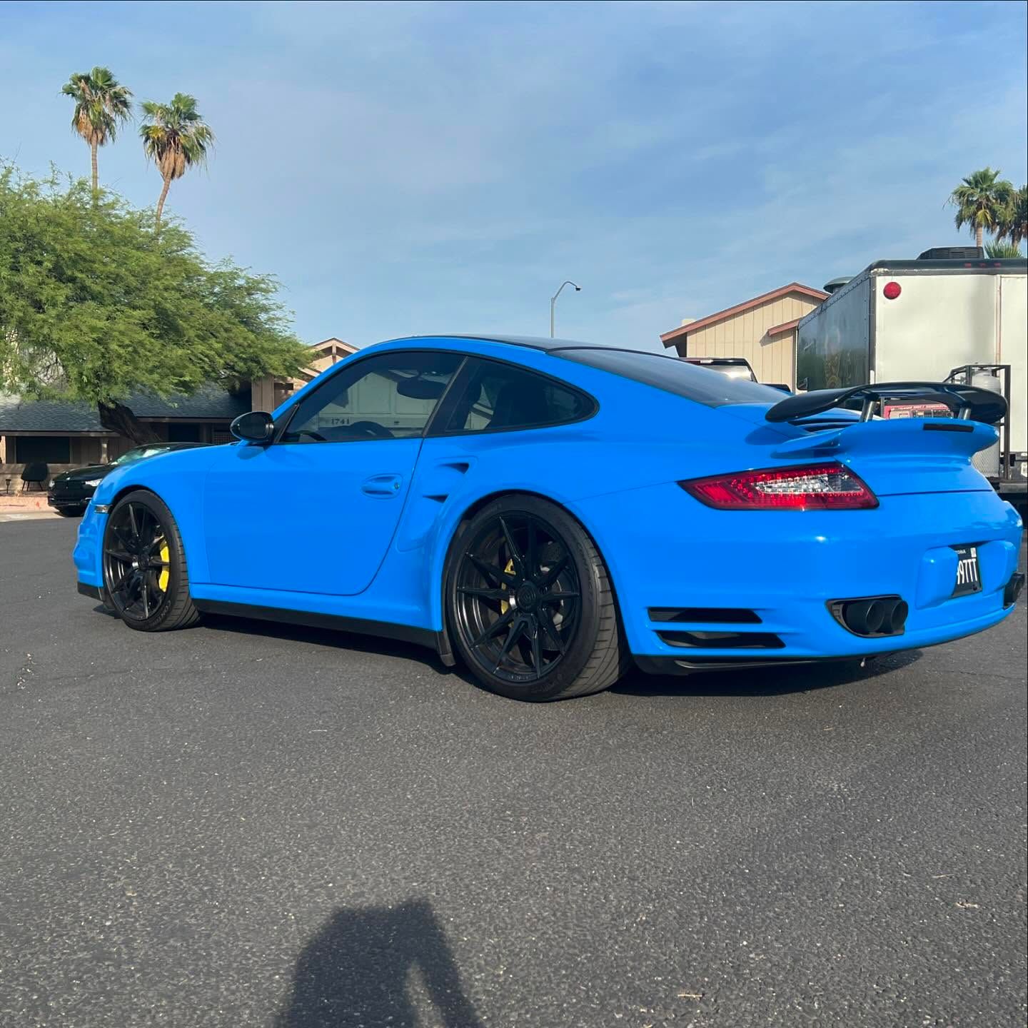 Bright blue Porsche sports car parked on asphalt. Black wheels, rear spoiler, palm trees in background.