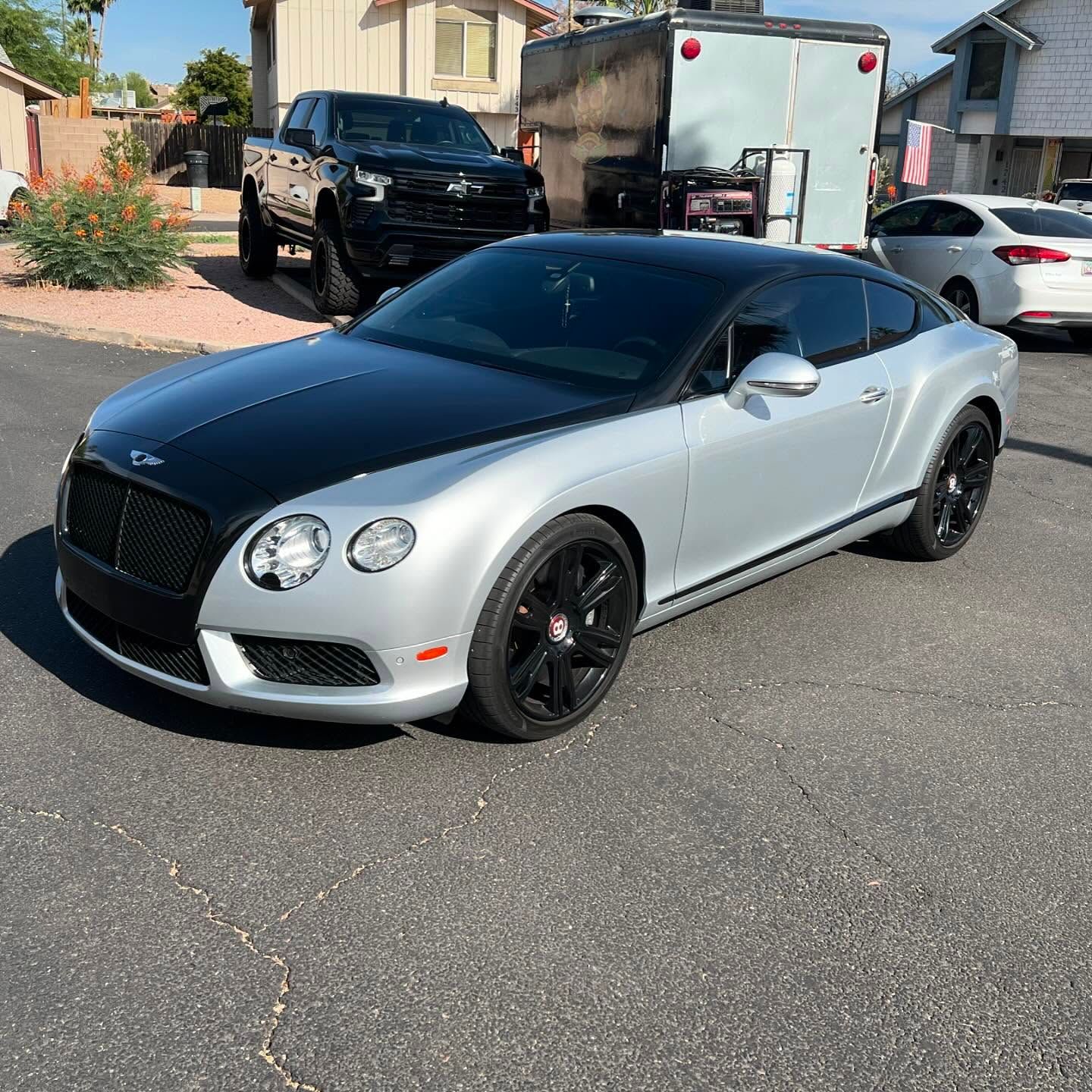 Silver and black Bentley coupe parked on asphalt, black wheels, black top, in front of a black truck and white trailer.