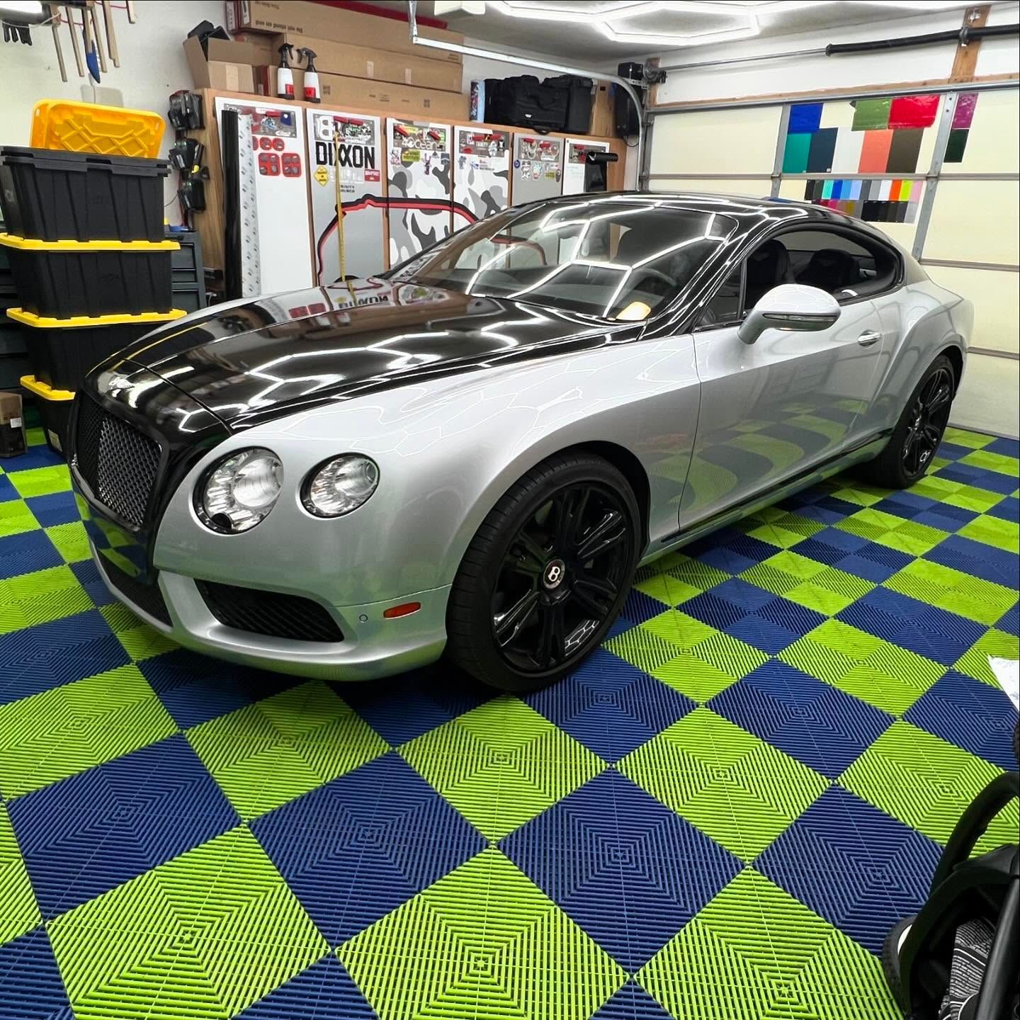 Silver Bentley coupe with a black hood in a garage on green and blue checkered flooring.