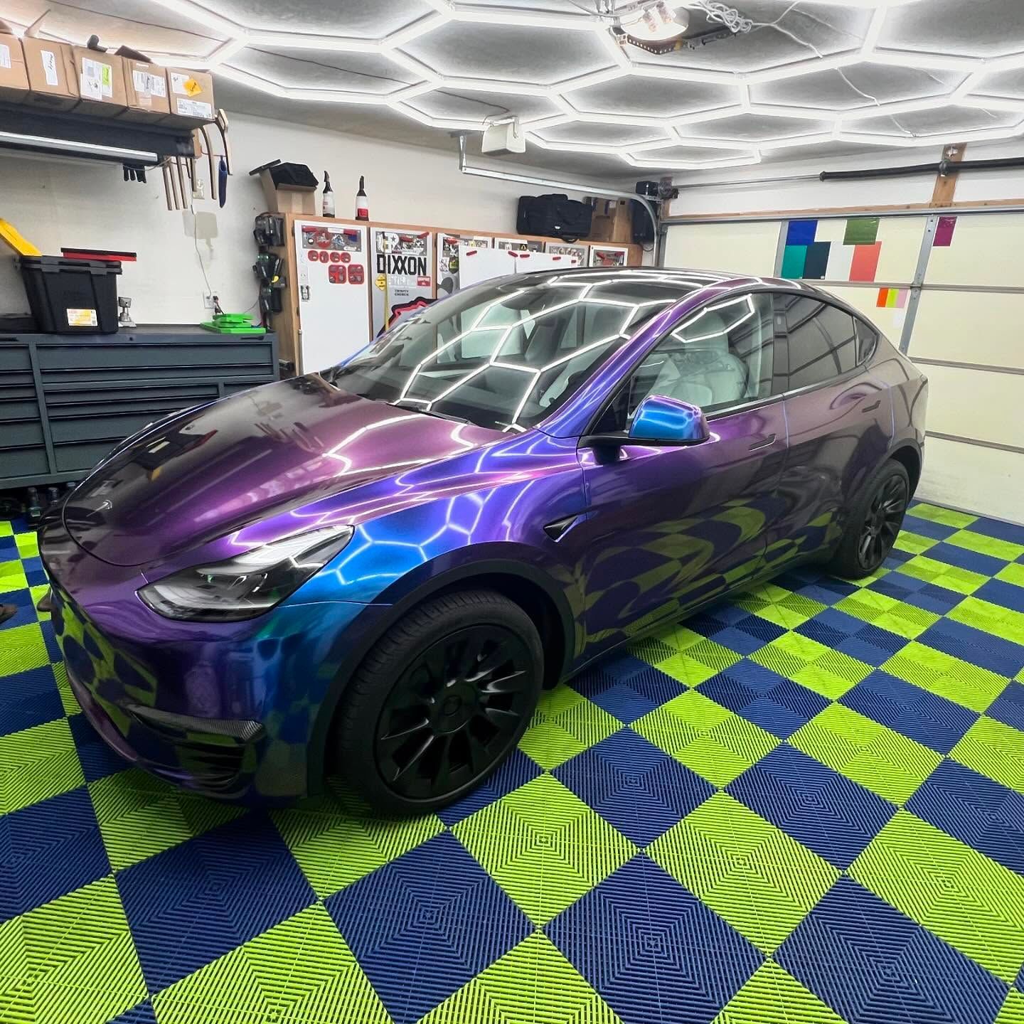 Tesla with a purple and blue iridescent wrap parked in a garage with hexagon lights and a checkered floor.