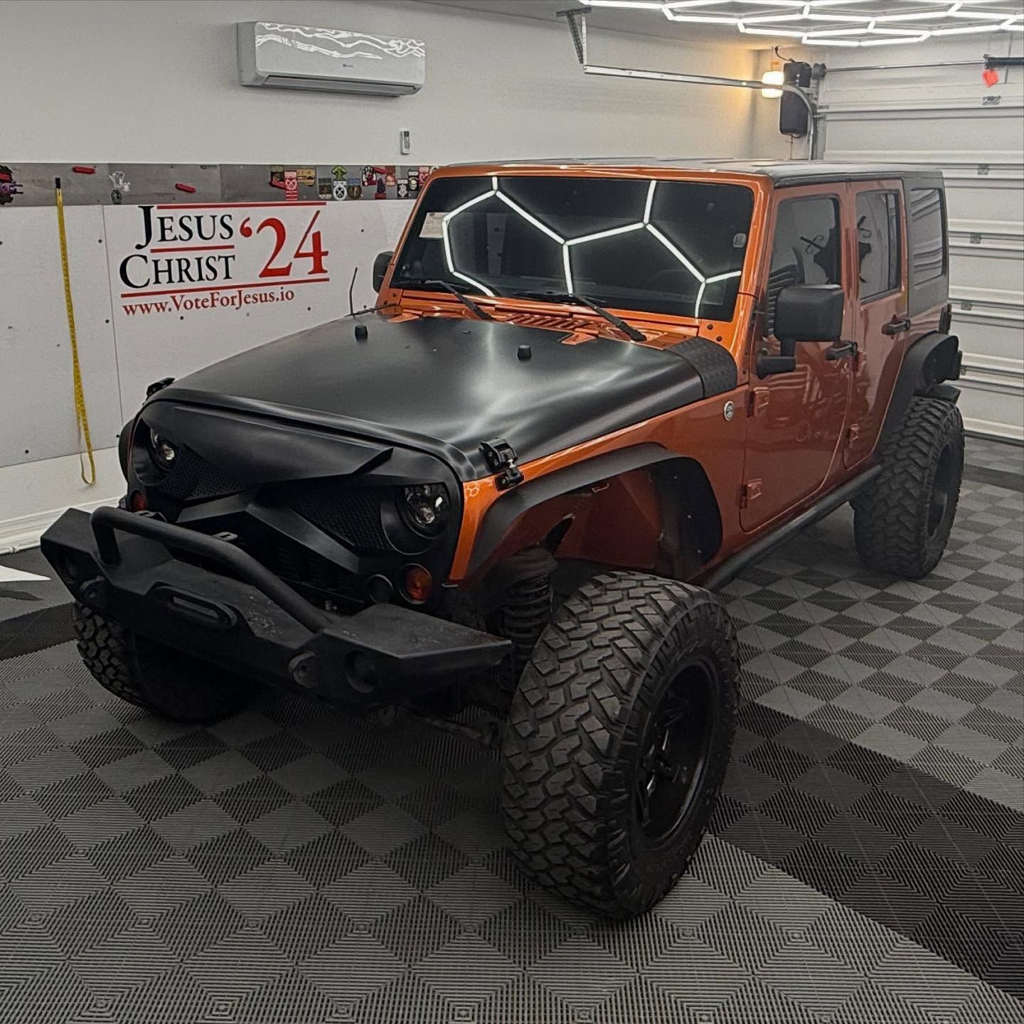 Orange Jeep Wrangler with black hood, bumper, and wheels in a garage.