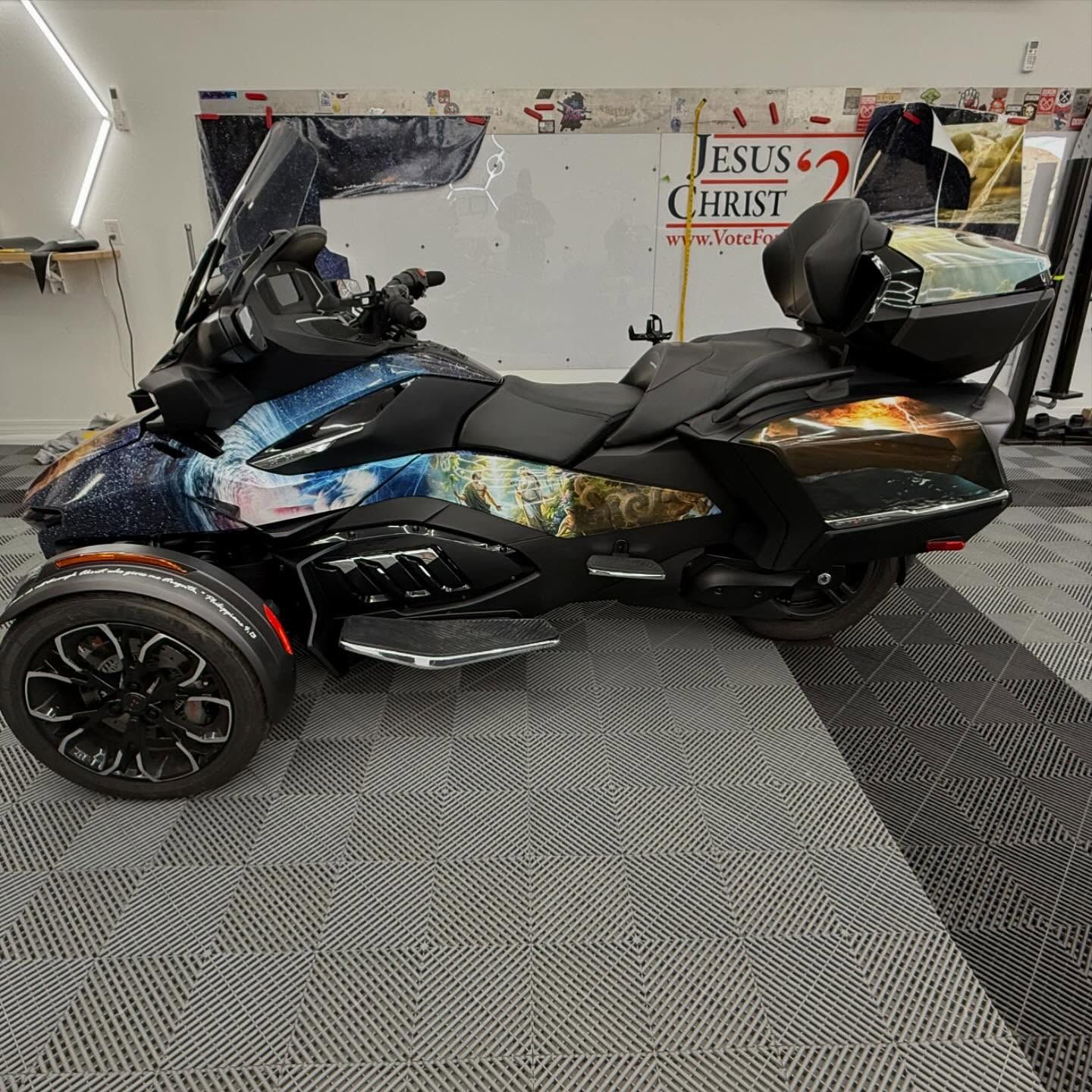 Three-wheeled motorcycle with colorful wrap; parked inside, near a white wall with religious text.