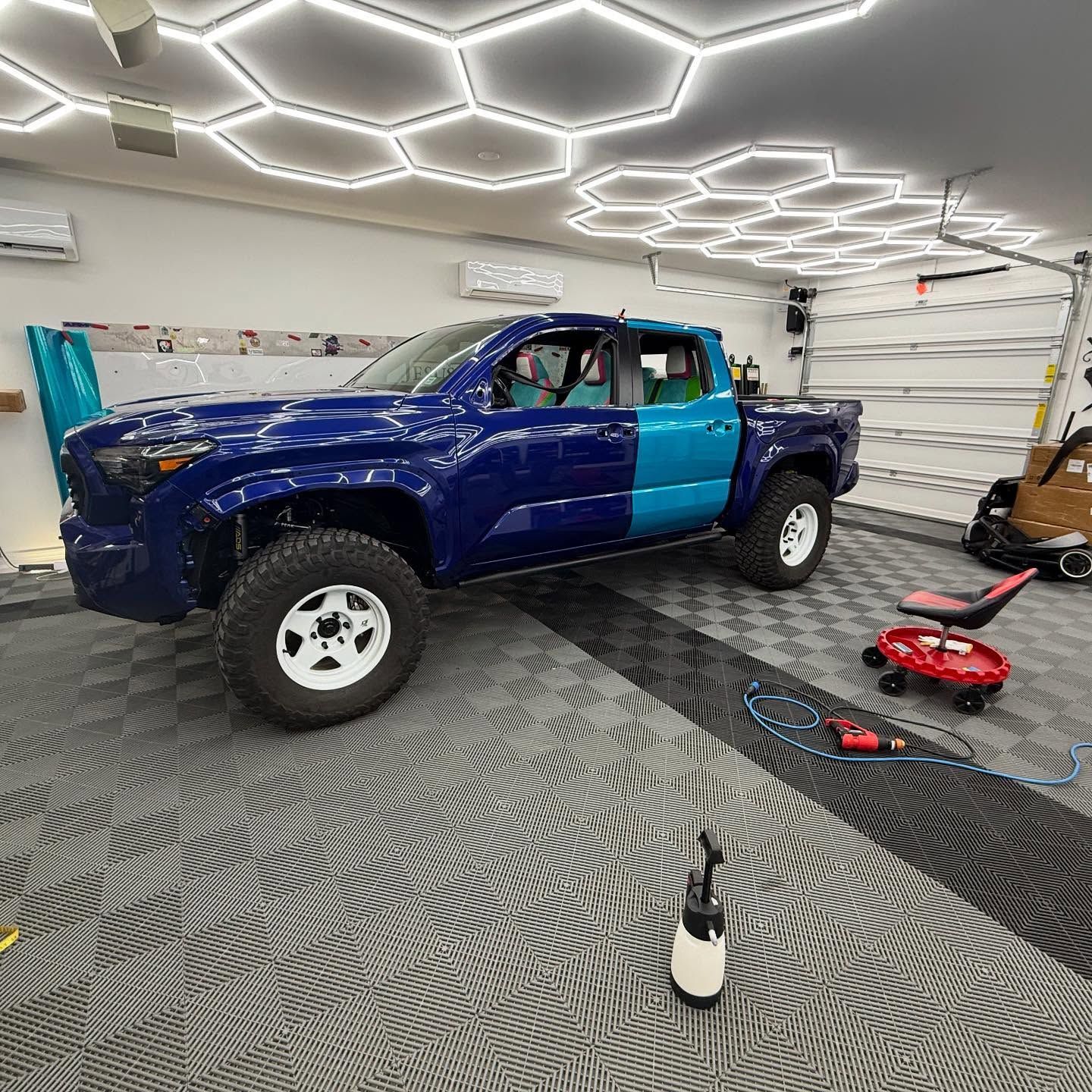 Two-tone blue pickup truck in a garage with white wheels. Garage has hexagon lights and tiled floor.