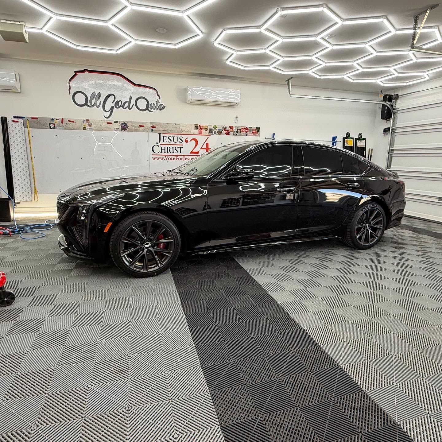 Black car in a well-lit garage. The garage has hexagon lights and tiled floors. The car has tinted windows.