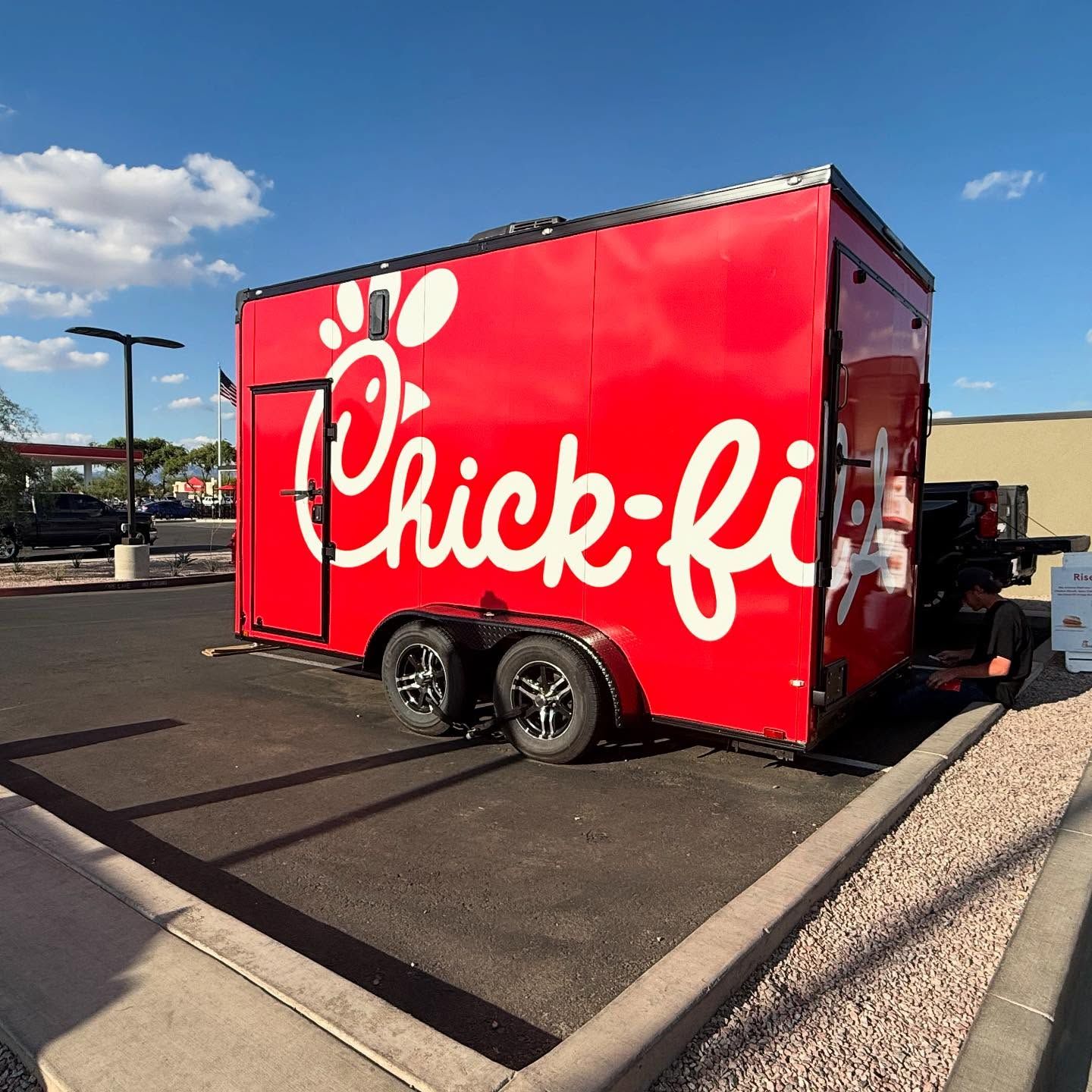 Red Chick-fil-A food trailer parked on asphalt, with a logo and black frame against a blue sky.