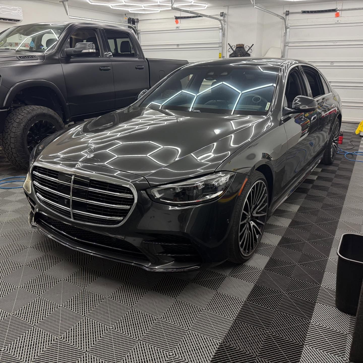 Black Mercedes sedan in a garage, next to a black truck, reflecting overhead lights.