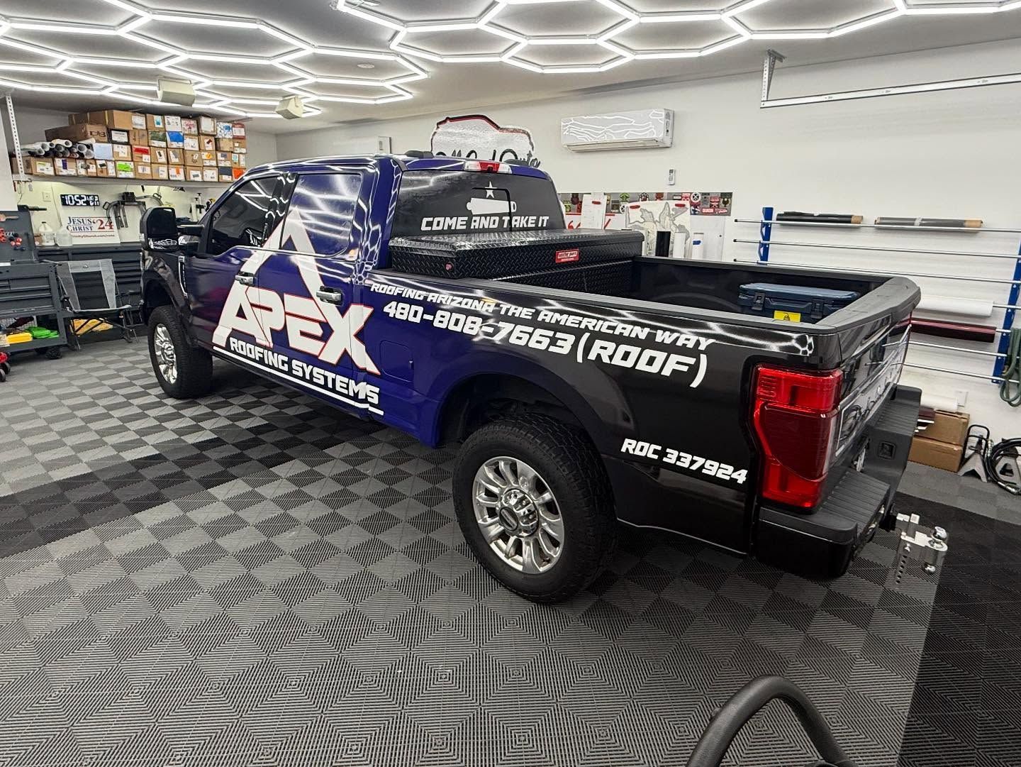 Blue and black Apex Roofing Systems truck in a shop, with the company logo and phone number visible.