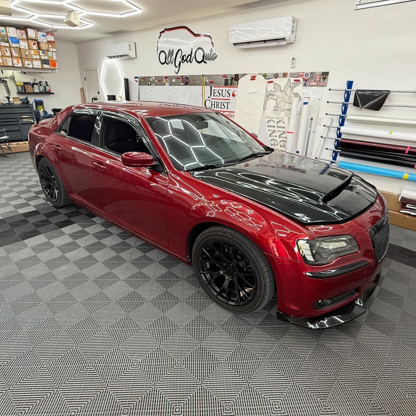 Red Chrysler 300 sedan with black hood, black wheels, and front spoiler in a shop with unique flooring.