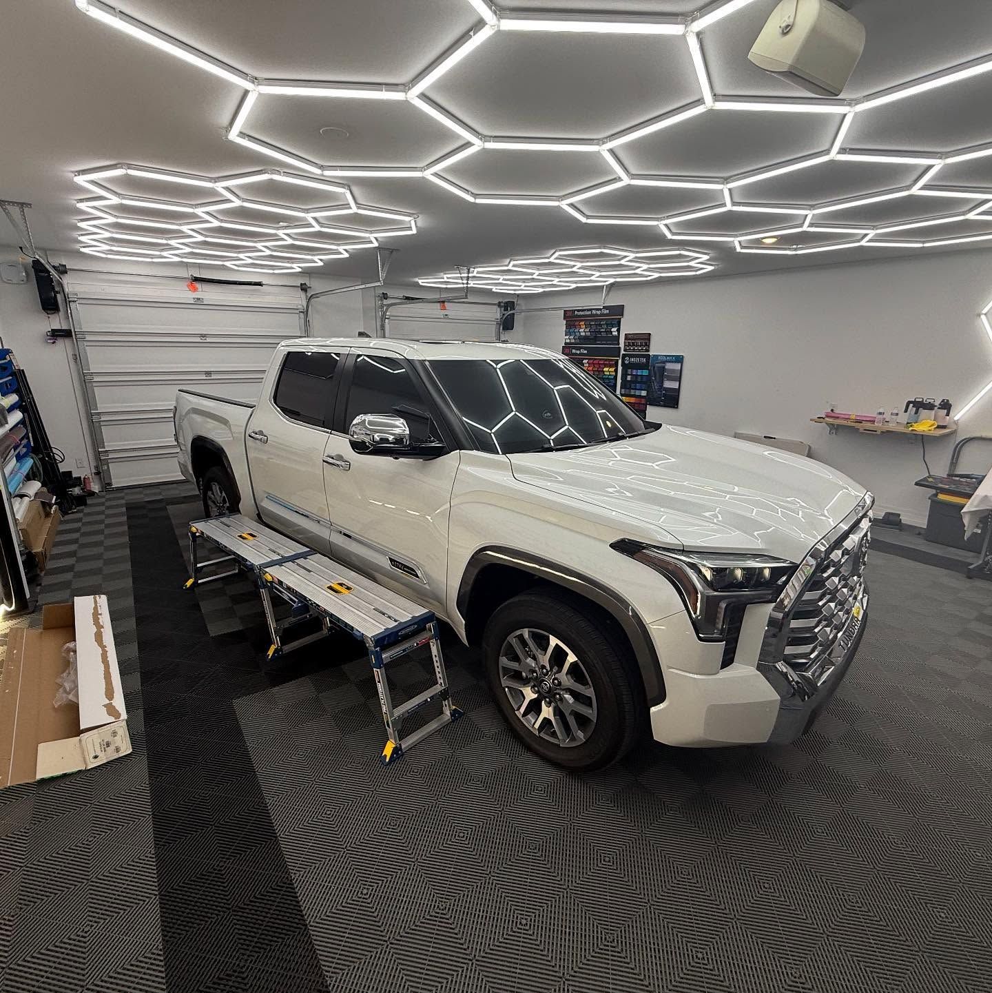 White truck on work platform in a brightly lit garage with honeycomb lights and black floor tiles.