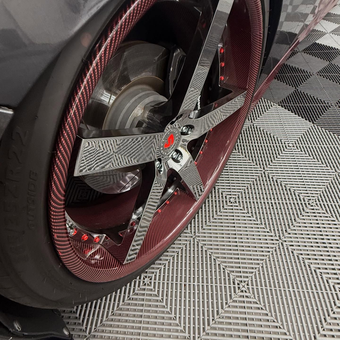 Car wheel with carbon fiber rim and chrome spokes, red accents, and a checkered floor.