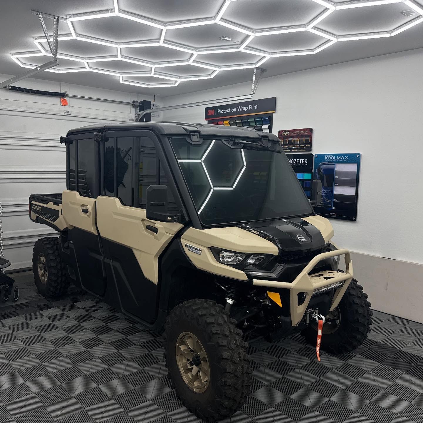 Beige and black Can-Am Defender UTV parked in a garage under honeycomb-shaped lights.