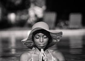 Black and white photo: Woman in a hat, in a pool, hands clasped near face, eyes closed.