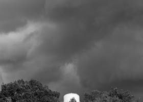 Dark, stormy clouds loom over trees and a white, cylindrical structure.