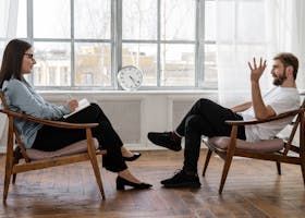 A therapist and patient in a therapy session, discussing. The setting is a room with a window.
