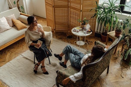 Two women in a therapy session: one seated, the other reclining on a couch in a bright room with plants.
