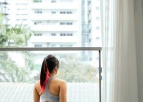 Woman with pink-streaked hair looks out a window. Building and trees in background.