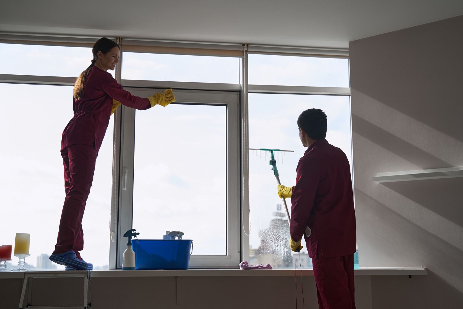 Two people in red jumpsuits cleaning a large window. One stands on a ladder, wiping the frame. The other uses a squeegee.