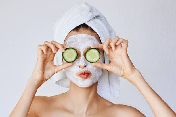 A person wearing a white face mask and hair towel holds cucumber slices over their eyes while making a playful expression.