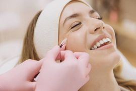A practitioner in pink gloves administers a facial injection to a smiling person wearing a headband in a clinic setting.