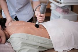 A technician uses an ultrasound therapy device on a patient's back covered in conducting gel in a clinical setting.