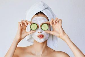 Person with a white face mask and towel wrap holding cucumber slices over their eyes.