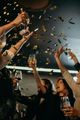 A group of people cheers with glasses raised as gold confetti falls in a dimly lit setting.