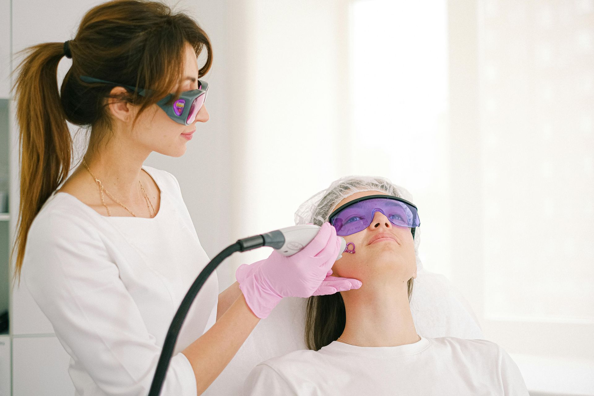 A clinician wearing pink gloves performs a facial light treatment on a reclining patient in a bright clinic.