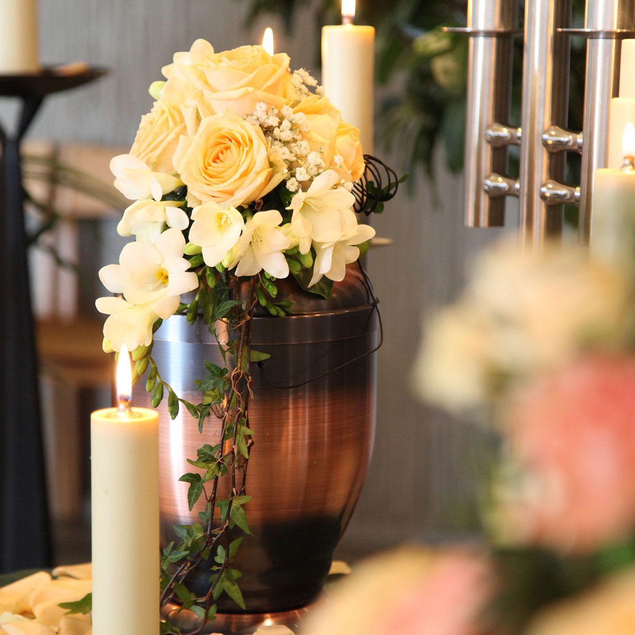 Copper cremation urn with floral arrangement and lit candles.