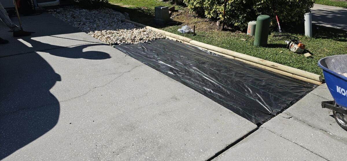 A person is laying a black tarp on a sidewalk next to a wheelbarrow.