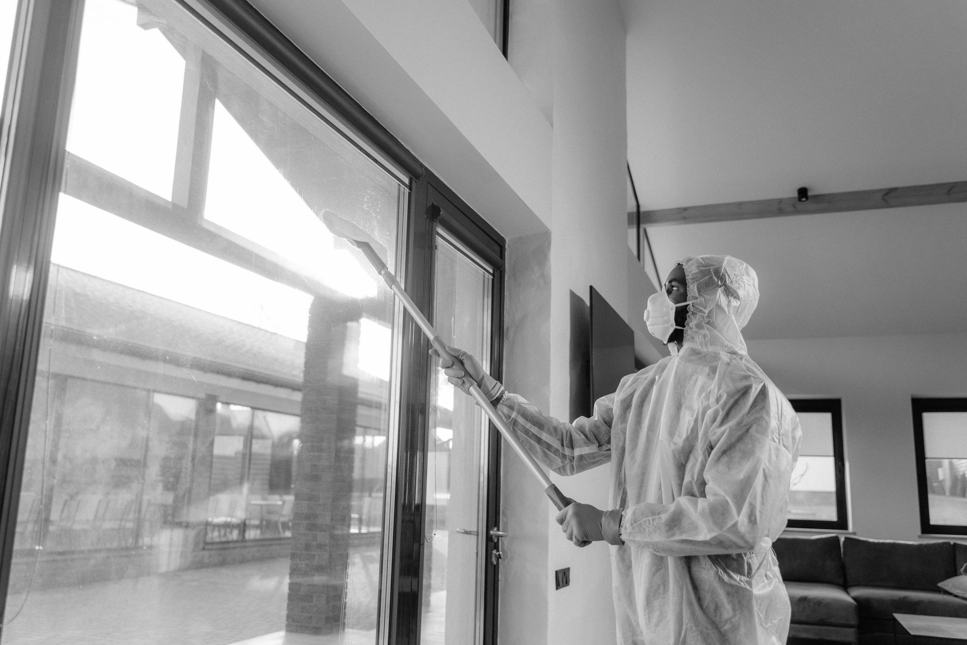 Person in hazmat suit cleaning a large window with a long-handled tool indoors.