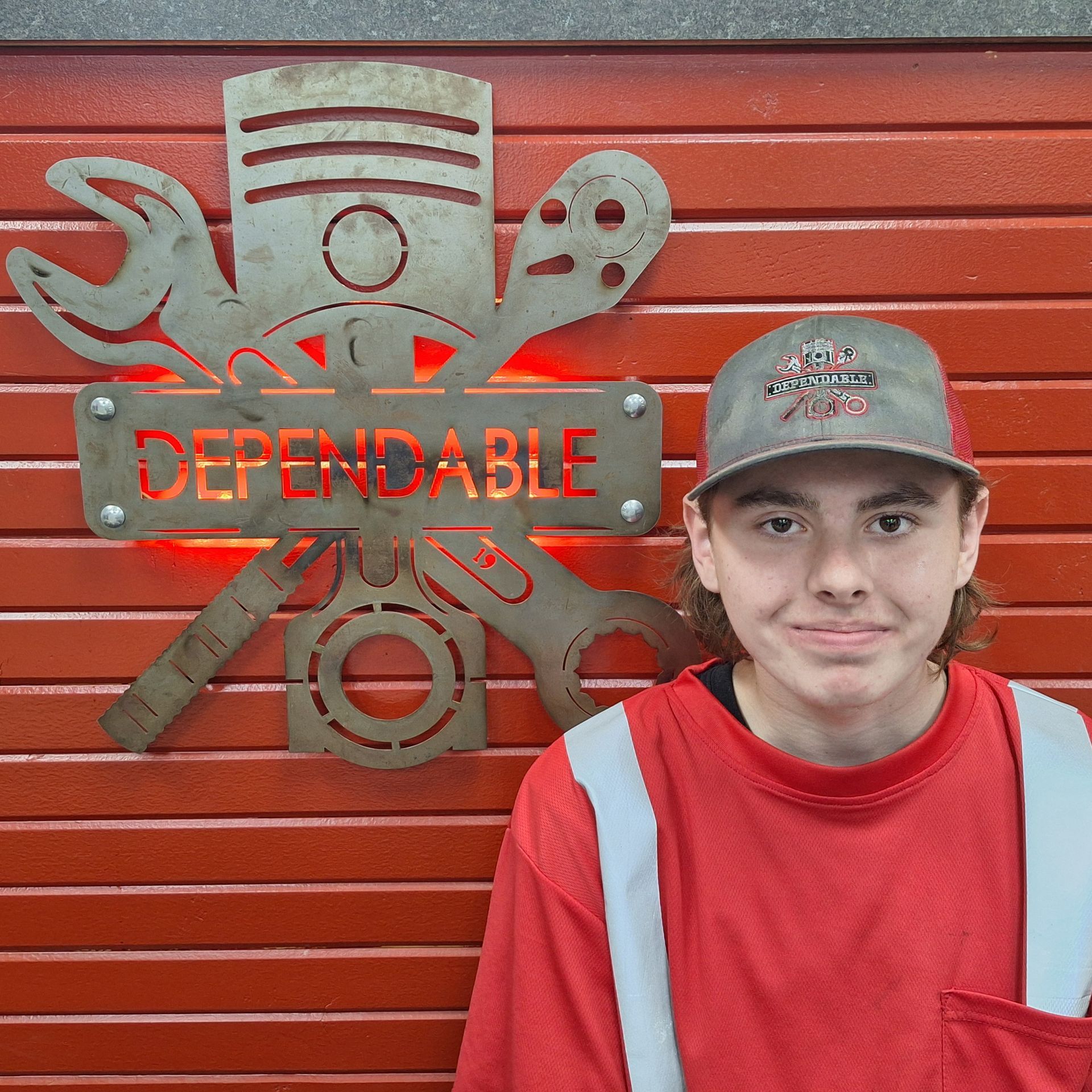 Young person in a blue work jacket and cap stands beside a red wall with a “Dependable” emblem. | Dependable Automotive