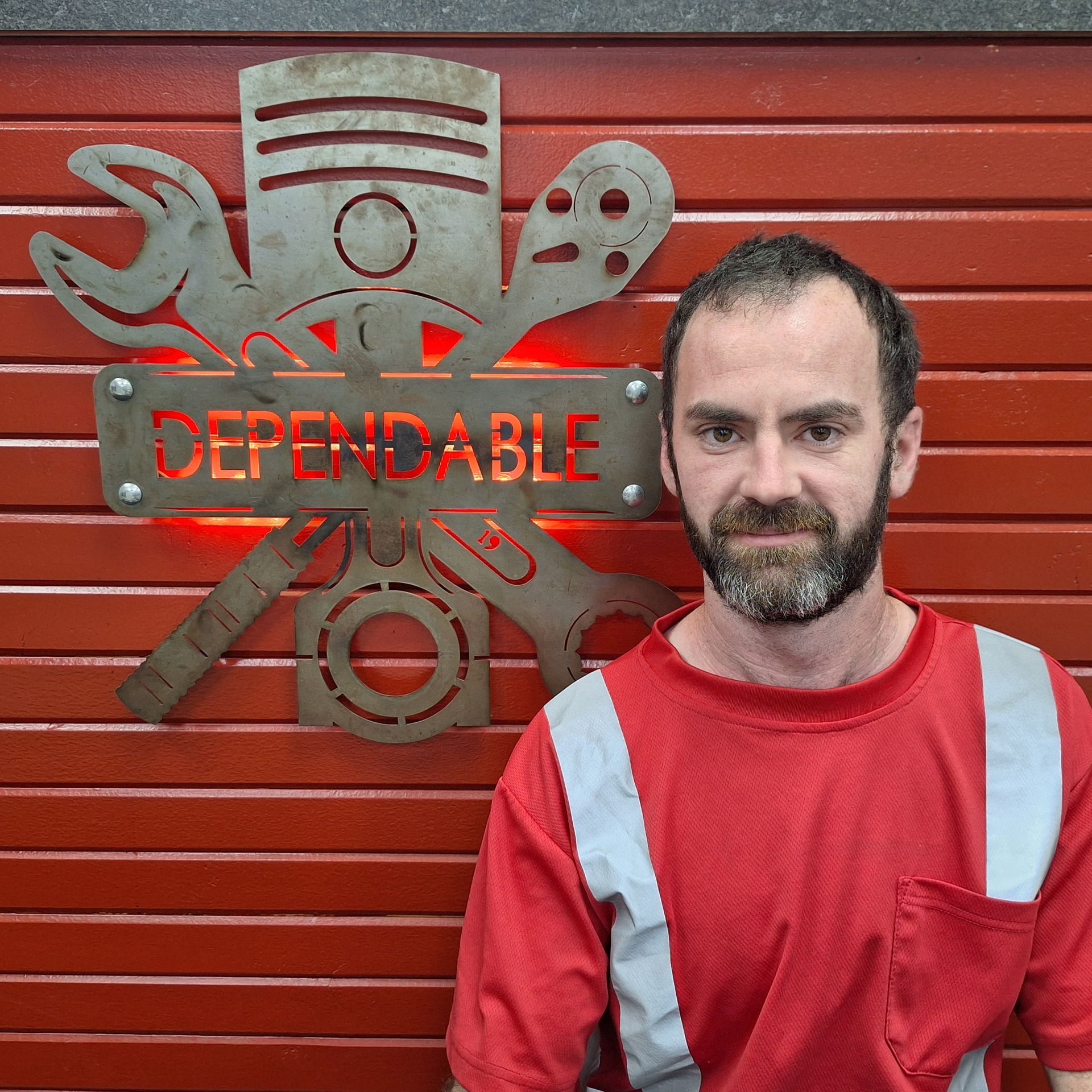 Smiling man in a black shirt standing before a red wall with a metal sign reading “DEPENDABLE.” | Dependable Automotive