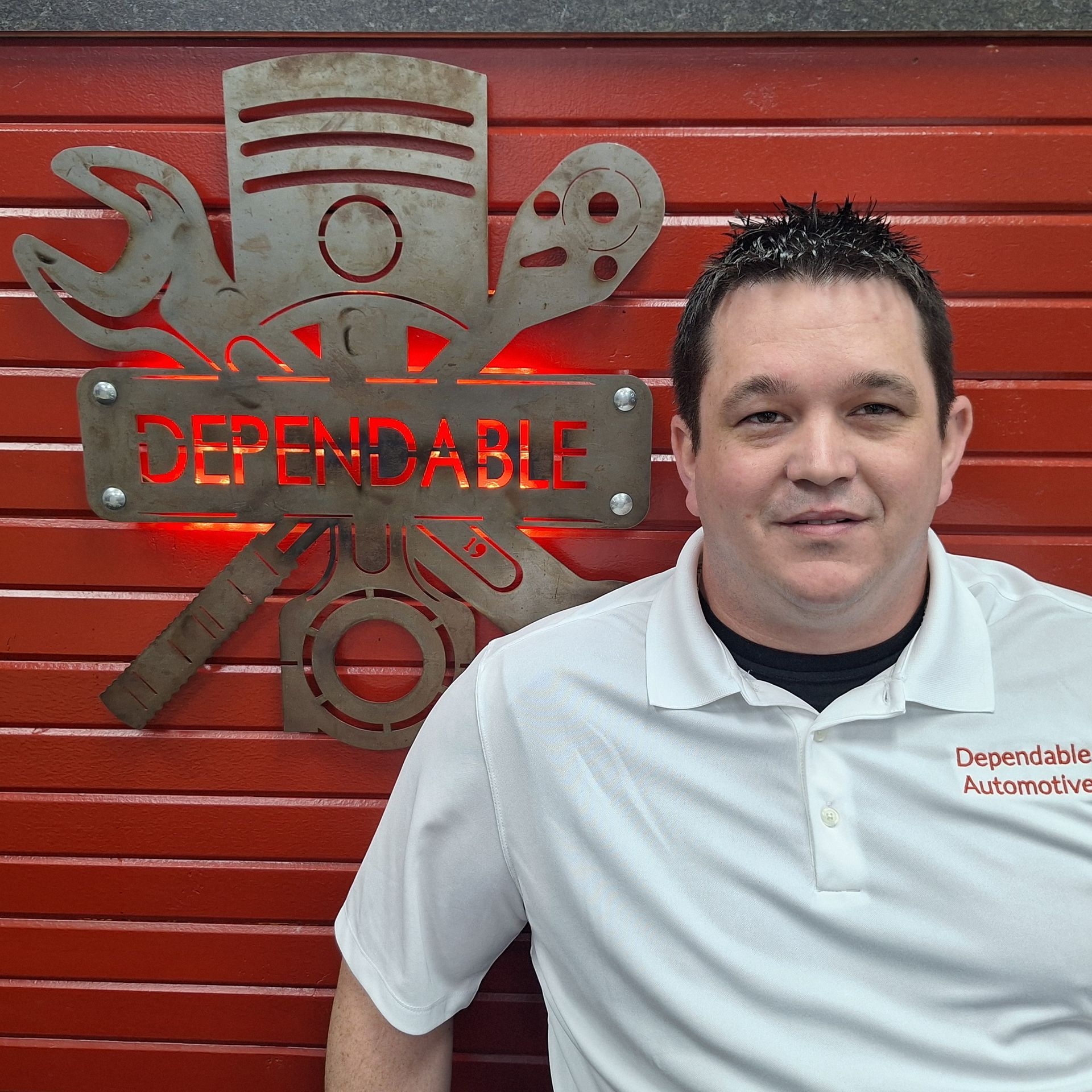 Man in a white shirt and cap stands before a red wall with a “Dependable” mechanic sign. | Dependable Automotive