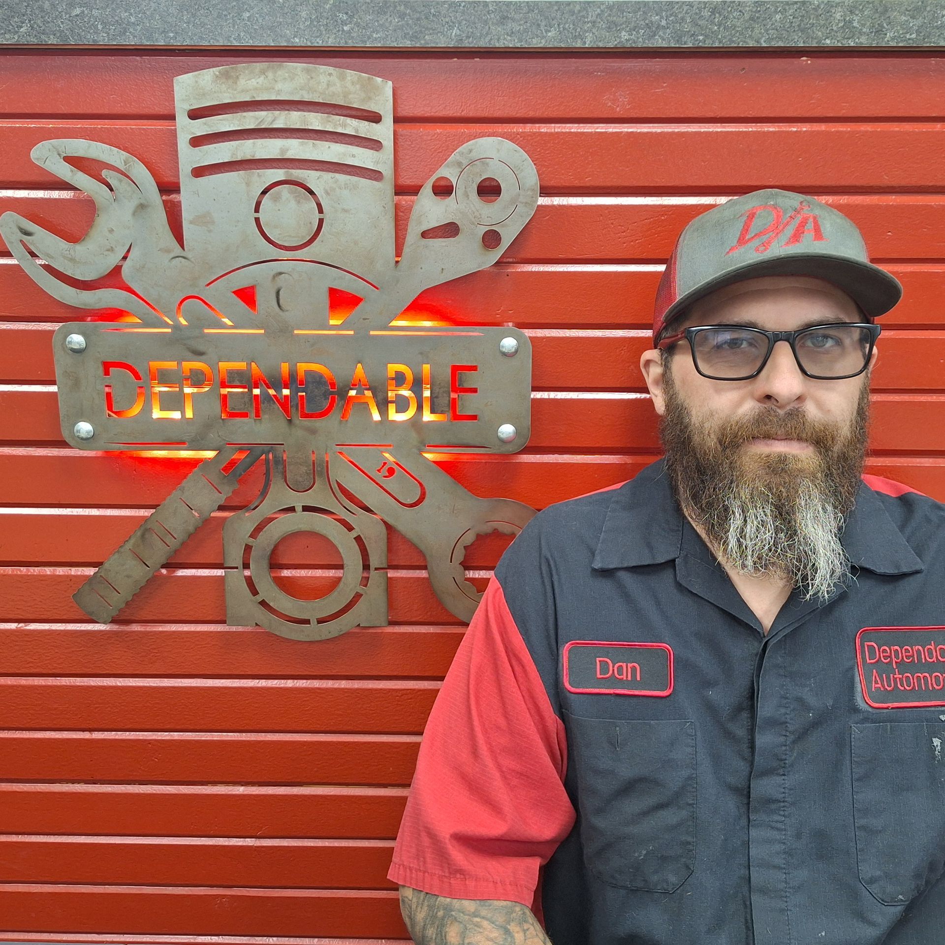 Smiling mechanic in red-and-black uniform and cap standing before a red Dependable sign. | Dependable Automotive