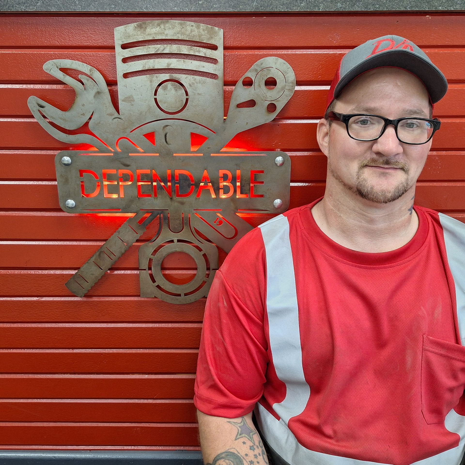 Young person in a blue work jacket and cap stands beside a red wall with a “Dependable” emblem. | Dependable Automotive