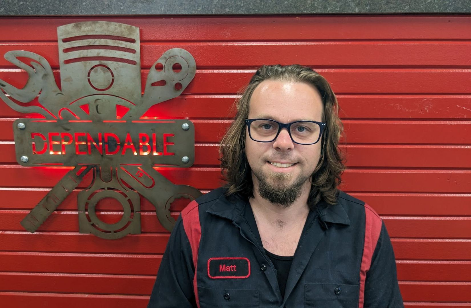 Man standing in front of a red wall beside a metal “DEFENDABLE” sign, wearing glasses and a black work shirt. | Dependable Automotive