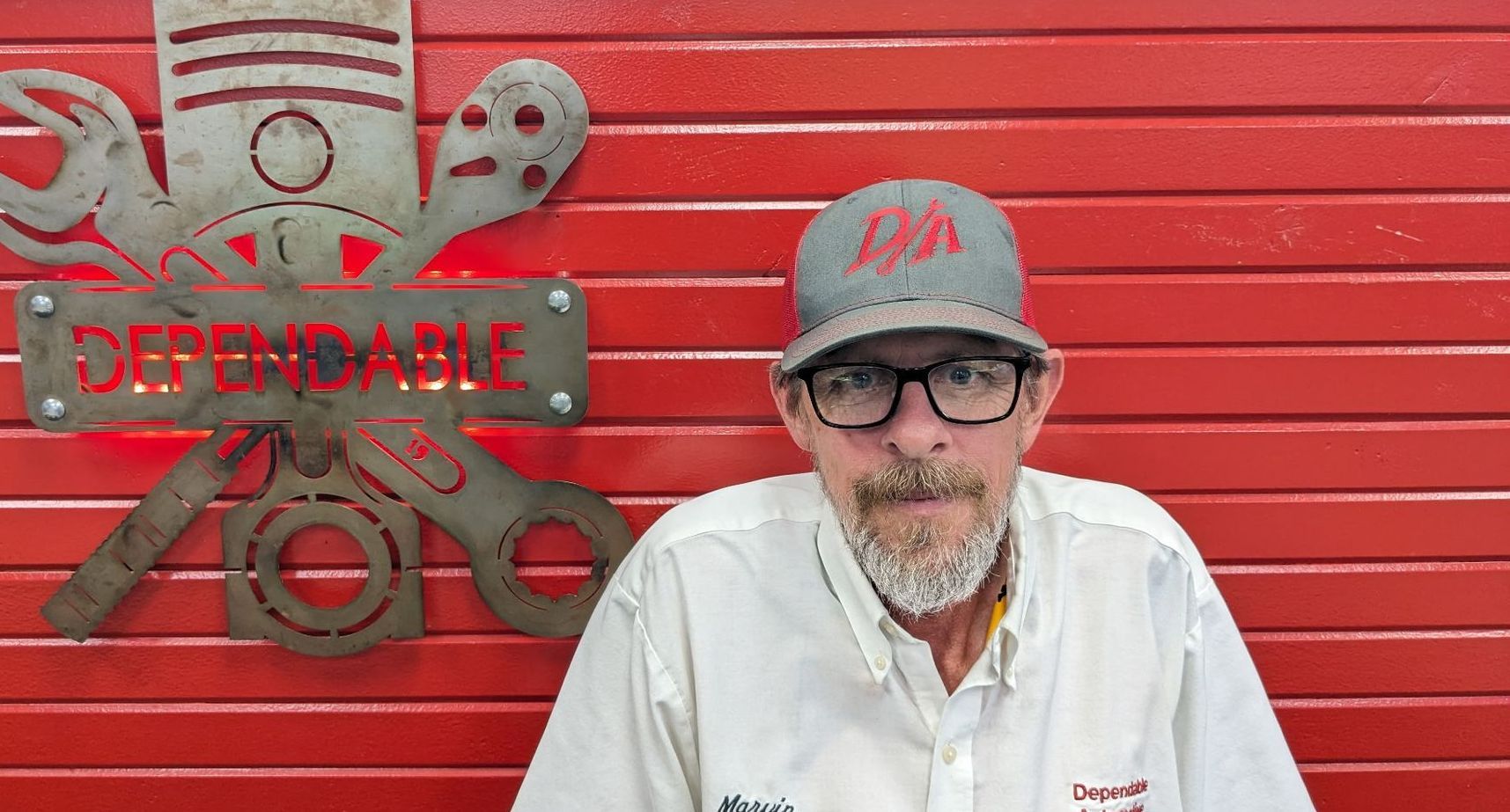 Man in glasses and red cap standing before red wall with a metal skull-and-crossbones sign | Dependable Automotive